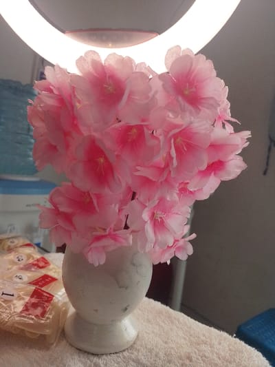 The scene is an indoor setting in Limuru Central ward, Kenya. A white, distressed, spherical vase with a pedestal base stands centrally, holding a dense arrangement of artificial pink fabric flowers. The flowers display lighter pink petals with darker pink centers and visible yellow stamens. The vase is positioned on a light-colored, textured fabric surface. To the left of the vase, multiple clear plastic bags containing light-colored artificial nail tips are present, each marked with red and white stickers featuring Chinese characters and numerical labels (e.g., "1", "2", "3"). An illuminated white ring light is partially visible in the upper background. The left background contains blurred light blue and white objects, while the right background is dark and out of focus. No individuals or actions are depicted.