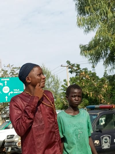In this medium shot, two individuals stand near a road in Garko, Nigeria. The person on the left, wearing a maroon traditional garment and a dark kufi, has their chin resting on their hand, with a contemplative expression. A string of wooden beads is draped around their neck. To their right, a younger individual in a green t-shirt with a faded graphic on the front looks directly at the camera with a neutral expression.

Behind them, a dark-colored vehicle, possibly a police car, is visible with red and blue lights on its roof. The vehicle has a circular emblem on its side with the word "NIGERIA" at the bottom. A green road sign is also present, indicating directions to "GOMBE" and "YOLA." The word "ABOUT" is visible within a circular arrow graphic on the sign.

The setting appears to be outdoors during the daytime, under a sky with scattered clouds. Trees with green foliage frame the upper right portion of the image. The overall impression is a candid street scene, possibly capturing a moment of pause or travel.