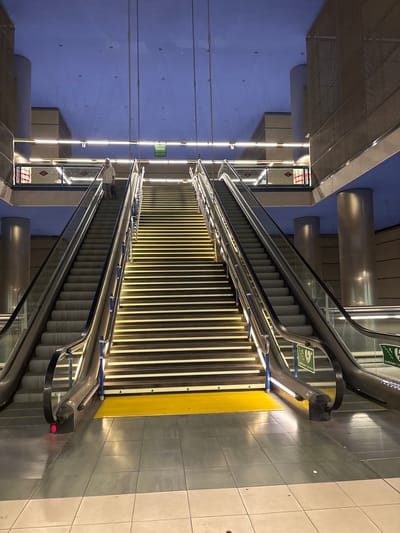 The image shows a long escalator in what appears to be a subway station in San Fernando de Henares, Spain. The escalator is illuminated by a series of warm-toned lights running along its sides, highlighting the metallic structure and the steps. A yellow strip runs along the edge of the escalator's landing area, likely for safety. The walls of the station are painted a cool blue, contrasting with the warm lighting. Tall, cylindrical columns are visible in the background.

On the right side of the escalator, there are green signs with pictograms that appear to indicate an emergency exit or safety information. There is also a sign with text that appears to say "Salida," which is Spanish for "Exit." On the left side of the escalator, there are what look like diamond-shaped signs with red and white markings, possibly indicating station branding or information.

A person wearing a light-colored shirt and dark pants is visible at the top of the escalator, standing and looking downwards. Another person is partially visible near the bottom of the escalator, also standing. The overall atmosphere seems to be one of transit and urban infrastructure, likely during the evening or night given the artificial lighting. The scene is clean and modern.