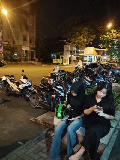 The scene takes place at night on a street in Telukjambe, Indonesia, at coordinates -6.308 latitude and 107.282 longitude.  There are several motorcycles parked along the street, facing a building in the background. Three individuals are present: two women sitting on a concrete planter and a man standing in the background. The women are both dressed in black, with one wearing a black hijab. One woman holds a green helmet in her hands and is looking at a mobile phone. The other woman is looking at a mobile phone. The man in the background appears to be looking at the motorcycles. The street is lined with small trees.  The scene is illuminated by streetlights.