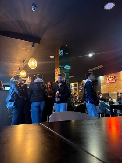 A dimly lit bar features several patrons. A group of people stand at a bar or table, dressed casually in jackets and jeans. Other customers are seated further back, including some in a booth-style setup. Above the patrons are various light fixtures, including hanging spherical lamps and recessed ceiling lights. A Red Bull neon sign is visible. The bar has wood paneling. A security camera is mounted on the wall. Sagres logos and branding are present on a structural element.