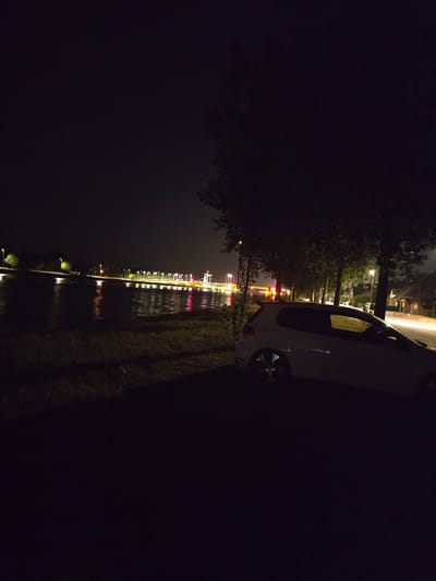 The image depicts a nighttime scene by a body of water, likely the canal in Zelzate, Belgium. A white Volkswagen Golf GTI is parked on the grassy bank to the right of the frame, its headlights off. The water reflects the lights from street lamps and structures along the opposite bank, creating a shimmering effect. Trees line the bank behind the car, casting shadows. In the background, streetlights illuminate a path or road, and some buildings are visible. The overall impression is one of a quiet, nocturnal urban environment.