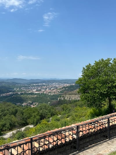 A bright, sunny day presents a panoramic view of the city of Arbanassi, Bulgaria, nestled in a valley surrounded by rolling green hills. The foreground is dominated by a rustic terracotta-tiled roof and a black wrought-iron fence, suggestive of a vantage point from a historical building or viewpoint. A lush, green deciduous tree stands prominently to the right, its branches reaching towards the clear blue sky dotted with sparse white clouds.
The city itself, appearing as a sprawl of buildings with red-tiled roofs and modern structures, lies in the mid-ground, bordered by dense forests. The terrain is varied, with visible cliffs and numerous trees covering the landscape. In the distance, layers of hazy mountains recede into the horizon under the expansive sky. A lone figure, small and indistinct, can be seen seated near the tree on the right, adding a touch of human presence to the serene scene. The overall impression is one of natural beauty and historical depth, characteristic of the Bulgarian countryside.