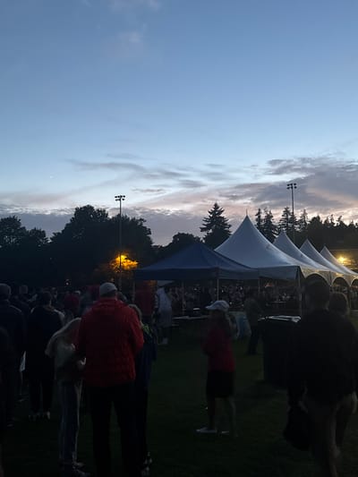 The image depicts a dusky outdoor gathering, likely an event or festival, taking place in New Westminster, Canada. A crowd of people stands on a grassy field, facing several large, white tents and a smaller blue tent in the background. The sky above is a gradient of pale blue and grey, with wisps of clouds catching the fading light, suggesting it is either dawn or dusk. A single bright streetlamp illuminates the scene from behind the trees on the left. The overall atmosphere is relaxed, with people milling about and some engaged in conversation. The setting appears to be a park or open field, possibly with athletic facilities suggested by the presence of a lamppost commonly found on sports fields.