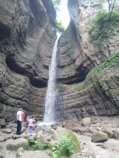 The image depicts a scenic view of a waterfall cascading down a rocky cliff face within a narrow canyon. The rock formations are layered and textured, with some areas covered in moss and vegetation. The waterfall itself is a prominent feature, falling from a considerable height into a shallow, rocky stream at the bottom. In the foreground, several people are visible, appearing to be tourists or visitors. Two adults and two children are standing on the rocky ground near the base of the waterfall, observing the natural scenery. The environment appears to be natural and somewhat rugged, with scattered rocks and foliage. The location is identified as Khushto-Syrt, Russia.