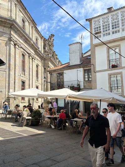 This media file captures a sunny day in Santiago de Compostela, Spain. The scene depicts an outdoor seating area of a cafe or restaurant, with several people seated at tables and others standing. The architecture suggests a historical European setting, with a stone building featuring arched windows and ornate decorative elements. A large white umbrella shades some of the tables, indicating a bright, sunny time of day. People are casually dressed, suggesting a relaxed atmosphere. Some individuals are engaged in conversation, while others appear to be dining or enjoying beverages. The image is oriented vertically, with the sky and buildings occupying the upper portion and the seating area and cobblestone ground in the lower portion.