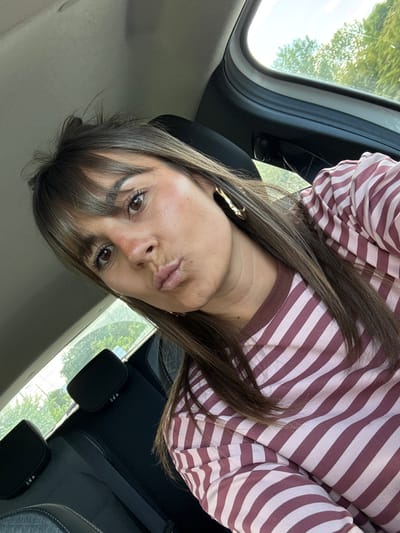 A woman with brown hair and bangs is seated in a car, wearing a pink and white striped shirt and gold earrings. She is looking directly at the camera with a slight pout. The car appears to be parked, with a view of trees visible through the window. The location, Les Arcs, France, is not directly visible in the image.