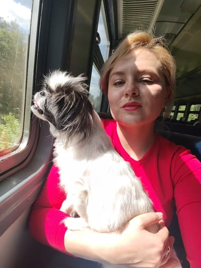 This is a selfie taken inside a train car, likely in Pugachyovo, Russia. The main subjects are a young woman with blonde hair tied back and a small black and white dog. The woman is wearing a bright red top and is holding the dog, which is looking out the window with its tongue slightly out. Sunlight streams in from the window, illuminating the woman's face and casting shadows. The background shows the interior of a train, with other passengers visible in the distance and the characteristic slatted ceiling. The window reveals a view of trees and greenery, suggesting a daytime journey. The overall mood is relaxed and casual, perhaps conveying a sense of travel and companionship. There is no visible text in the image.