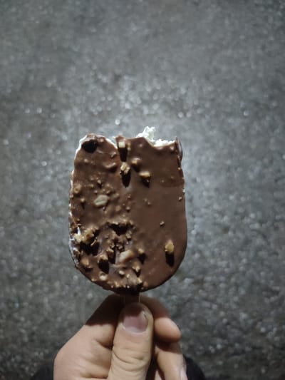 The image displays a single human hand holding a partially consumed chocolate-coated ice cream bar with visible nut pieces, set against a dark, out-of-focus, granular background, in Rishtan, Uzbekistan. The ice cream, positioned centrally, shows a distinct bite taken from its upper-left side, exposing the white frozen confection underneath the melted-appearing chocolate shell. A light-skinned hand, exhibiting the thumb and index finger, grips the wooden stick of the ice cream. The background, characterized by blurred light reflections, suggests an outdoor or dimly lit environment with a textured surface like asphalt or concrete.