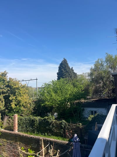 The image shows a view from an elevated position, possibly a balcony, overlooking a garden and the surrounding environment in Maastricht, Netherlands. The foreground includes a brick wall and dense foliage of various trees and bushes. A partial view of a building with a white exterior and overgrown ivy is visible. Elements of outdoor furniture and garden implements are also present. In the distance, there are railway tracks and electrical pylons. The sky is blue with scattered clouds, indicating a clear day. An antenna is visible in the top left corner.