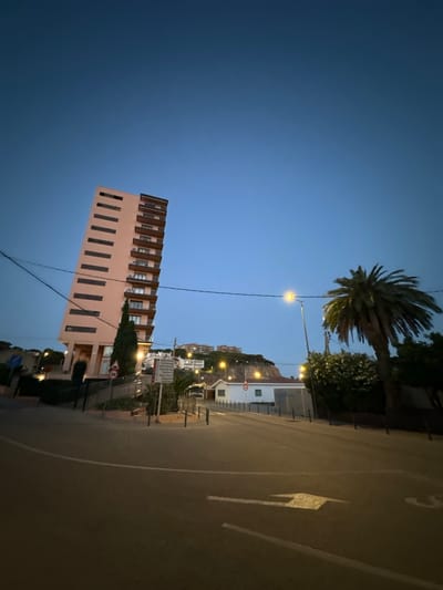 The image captures a twilight scene in Sant Feliu De Guíxols, Spain. A tall, slender, peach-colored apartment building with horizontal dark-framed windows and balconies dominates the left side of the frame. In the background, a palm tree stands to the right of the apartment building, with other greenery and buildings on a hillside. A streetlamp illuminates the area, casting a warm glow on the surrounding road and pavement, which features white lane markings, including an arrow indicating direction. The sky is a deep blue, transitioning from the darkness of night. Several power lines are visible stretching across the sky. A white rectangular sign with text and a speed limit sign indicating "20" are placed on the side of the road. A single-story white building with red trim is visible on the right side of the frame, near the palm tree. The overall impression is one of a quiet, possibly residential, urban environment at the end of the day.