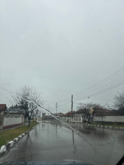 The image shows a view from inside a vehicle, likely a car, looking out through a rain-streaked windshield. The scene is a residential street in what appears to be a cloudy or overcast day. The street is wet, reflecting the light and the shapes of buildings and trees. Houses with fences and greenery line the street. Electrical poles and wires are visible overhead. A street sign is present near a house. A curb with white and dark road markings is visible along the grass lawn. The location of this scene is near Poroino, Bulgaria.