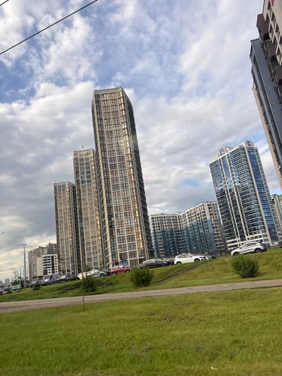 The image captures a contemporary urban landscape with a series of high-rise apartment buildings in the distance. The buildings exhibit modern architectural styles, featuring multiple stories, numerous windows, and varying facade designs, with some boasting glass balconies and intricate patterns. Several cars are parked along the side of the road and on an elevated grassy slope in the foreground, with a green tram also visible. The sky is partly cloudy, with a mix of white and gray clouds against a blue backdrop. The foreground is dominated by a lush green lawn, suggesting a park or roadside green space. The overall scene conveys a sense of a developing residential area within a city, likely in the Сеніцкі сельскі Савет, Belarus, given the context.