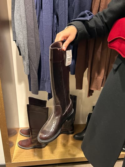 The image, taken in Chalkida, Greece, depicts a close-up, rotated view inside what appears to be a retail store, likely a clothing and accessories boutique. A person's hand, adorned with a silver bracelet, is holding a dark brown or burgundy knee-high boot by its shaft. The boot has a glossy, possibly patent leather, finish. A white tag, clearly visible on the boot, displays "Melvin & Hamilton" and "SIZE 38", along with a barcode and other unreadable text.
The boot being held is part of a display. To its right, several other boots are visible on light-colored shelves with a distinct golden or bronze-colored trim. Another pair of dark brown/black boots stands on a shelf directly below the held boot, and a pair of dark red-brown boots are further to the right.
Behind the hand and the boot, neatly folded garments are stacked on shelves. These include piles of brown/camel-colored fabric, dark blue fabric (possibly trousers or jeans), and various shades of grey fabric, indicating trousers or skirts of different textures.
In the top left corner, a portion of a red bag or clutch is visible, next to the person's arm, which is clad in a dark-colored (likely black) long-sleeved garment, possibly a coat or jacket. A black garment and a black shoe are also partially visible in the top right quadrant of the image.
The scene captures a moment of shopping or merchandise examination in an indoor setting, with artificial lighting illuminating the display.