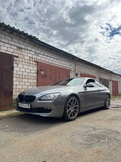 A gray BMW coupe is parked in front of a row of garages with brick walls and corrugated metal roofs under a cloudy sky. The car is facing the viewer at an angle, with its front license plate visible. The garages have large, rusty metal doors, some with numbers stenciled on them, such as "500". The ground in front of the garages is paved with asphalt and shows some water puddles. The scene appears to be in an industrial or garage complex.