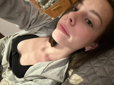 A young woman with fair skin and brown hair lies on a patterned gray surface, likely a bed or couch. She wears a black top and a gray zip-up hoodie. Her head is tilted towards the camera, showcasing her face with visible details like eyebrows, eyes, nose, and lips. Part of a wooden object is seen in the background. The image has been rotated, positioning her at an angle.