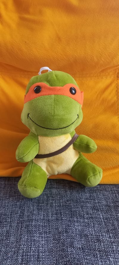 A stuffed toy, resembling a Teenage Mutant Ninja Turtle, is positioned centrally on a textured gray surface, likely a couch or chair. The toy is predominantly green, with a yellow plastron and an orange mask covering its eyes. A thin brown strap is visible across the toy's chest. Behind the toy, a bright orange fabric, possibly a blanket or throw, is draped. The lighting suggests an indoor environment, with soft illumination. There is no discernible location in the city of Идрисово, Russia associated with this image.