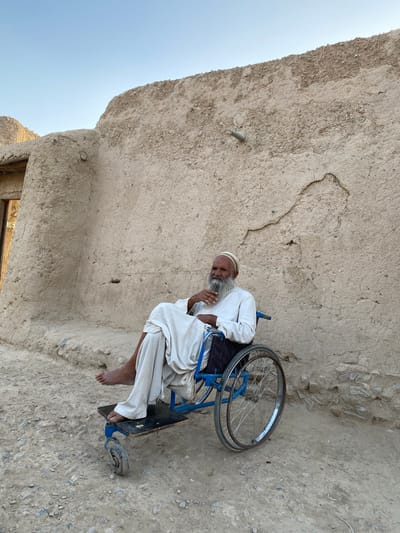 A man with a long, grey beard and a white skullcap sits in a blue wheelchair against a textured, mud-brick wall. He is wearing a white robe and appears to be resting, with his head tilted down and his hand near his face. His feet are bare. The wheelchair has large spoked wheels and a smaller caster wheel at the front. The ground is a rough, stony surface. The wall appears weathered and aged, with visible cracks and imperfections. The scene is outdoors, under a clear, pale blue sky, suggesting a daytime setting. The overall impression is one of quiet stillness. The location cues, such as the mud-brick architecture and the rough terrain, are consistent with a rural or traditional setting in Afghanistan. There is no visible text in the image.
