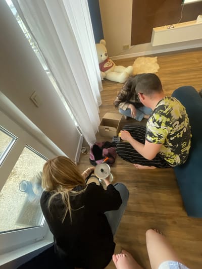 The image depicts an indoor scene, possibly a living room, featuring three individuals and various objects. The location is unspecified and not suggested in the image.
One person, wearing a yellow and black patterned shirt and striped pants, is kneeling near an open cardboard box. Another person with long hair is also kneeling nearby, possibly interacting with the contents of the box. A third person's bare feet are visible on the right side of the frame.
Objects in the scene include a teddy bear, a white sheepskin rug, a blue cushion or couch, a purple bag, and a cooking pot. There is a window in the lower part of the frame, with white curtains hanging. The floor appears to be made of wooden planks.