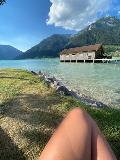 The image depicts a scenic view of a calm, clear lake bordered by lush green mountains and a blue sky with scattered clouds. In the foreground, a person's legs are visible, suggesting they are sitting on the grassy shore. The person appears relaxed and enjoying the scenery.

A wooden structure, possibly a boathouse, stands on stilts in the lake, with its reflection visible in the water. The water itself is transparent, allowing a view of the rocky bottom. The surrounding mountains are densely covered with trees, creating a beautiful natural backdrop. The lighting suggests it is daytime, likely late afternoon or early evening, given the warm glow on the grass and the shadows cast. The weather appears to be pleasant and sunny, ideal for outdoor activities.

The location, Lärchenwiese, Austria, further enhances the scene's natural beauty, known for its stunning alpine landscapes. The overall mood is serene and peaceful, capturing a moment of tranquility by the water. There is no visible text in the image.