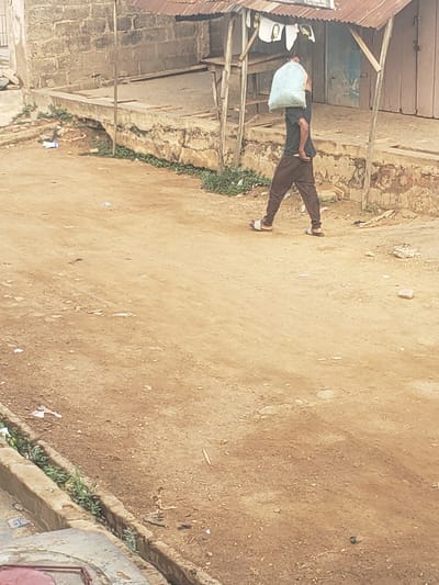 The image shows a man carrying a large light-colored bag over his shoulder, walking on a dirt ground in Alalubosa, Nigeria. He is wearing a dark shirt, brown pants, and sandals. The environment includes a building with a corrugated metal roof and brick walls on one side of the path, and a drainage ditch with vegetation on the other side. The ground is dry and dusty, with scattered debris.