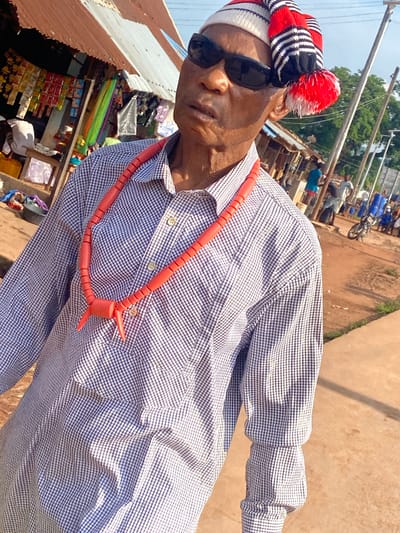 An elderly individual, likely male, is captured in a close-up, slightly upward-angled shot on a bright day in Amaseri, Nigeria. He wears dark sunglasses and a distinctive knitted cap featuring red, white, and black stripes with a large red and white pom-pom on the right side. A long, orange-red beaded necklace, ending in two pointed segments, rests over a long-sleeved, light-colored gingham shirt. His expression is directed slightly upward and to his left.
The background reveals a bustling outdoor environment. To the left, a rudimentary shop or stall with a rusty corrugated metal roof displays various packaged goods, including snacks and brightly colored fabrics. A person in white is partially visible behind the counter. To the right, an unpaved reddish-brown path extends into the mid-ground where several individuals are visible, some standing, some seated. A motorcycle is parked on the dirt path near some blue barrels. Utility poles and power lines traverse the clear sky, and distant trees mark the horizon under bright sunlight.