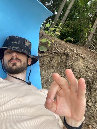 A man wearing a camouflage bucket hat and a light-colored ribbed tank top is lying on a bright blue surface outdoors. He has a beard and mustache and is looking towards the camera, with his hat pulled down over his eyes. His right hand is raised in front of him, with his thumb and index finger forming a circle, and his other fingers extended, in what appears to be an "okay" gesture. The background shows a rough, earthy embankment covered in roots and some greenery, with trees and foliage visible further up. The overall setting suggests an outdoor recreational area, possibly near a body of water given the potential for a beach or lakeside environment. This scene could be located in Kaniów, Poland, a town known for its natural beauty and proximity to the Vistula River, which offers opportunities for outdoor activities.