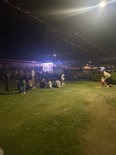 The image shows a group of people gathered in a grassy area in front of a stage, located at coordinates 38.433, 27.137, in İzmir, Türkiye. The stage is brightly lit, and there are several strings of red and white flags hanging above it. The people are standing and sitting on the grass, some are interacting, and one woman is walking with a young child. The scene is illuminated by streetlights and spotlights. There are trees and bushes surrounding the grassy area, and the background is a blurred image of buildings and trees. The image was taken at night.