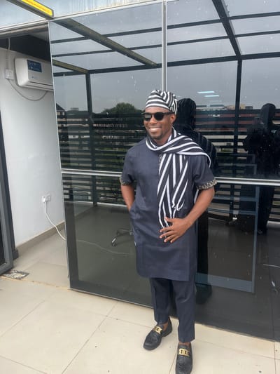 A man is standing outside a building, possibly in Karum, Nigeria. He is wearing a dark blue traditional outfit with a striped scarf and a matching headscarf. The outfit appears to be made of a light fabric, suitable for warm weather. He is also wearing dark sunglasses and black loafers with gold accents. The man is smiling broadly, suggesting he is happy or in good spirits.
The background features a modern building with large, dark glass windows and a black metal frame. The windows reflect the sky, which appears overcast, indicating it might be a cloudy day. The man is standing on a tiled floor, possibly a balcony or patio. An air conditioning unit is visible mounted on the wall below the windows. A white electrical cord is running from a wall socket to the unit. There is also some text visible on a yellow sticker on the far left side of the image. The overall impression is that of a casually posed portrait taken outdoors.