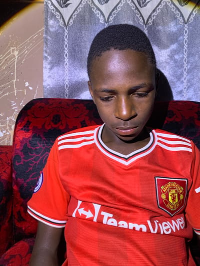 The media file shows a young Black person lying down, wearing a red Manchester United jersey with white stripes on the sleeves and "TeamViewer" text. The person's eyes are closed, and they appear to be relaxed or sleeping. They are lying on a surface that has a gray, patterned fabric, possibly a sofa or bed, with some red patterned fabric visible to the right. The lighting suggests it is indoors, possibly in the evening or a dimly lit room. The image's orientation is rotated, with the person's head to the left and their body extending towards the right. The background includes a patterned fabric with red and black floral designs and a beige surface with white paint splatters. The location is Barakin Sabawa, Nigeria. There are no specific cues for time of day or weather. The visible text on the jersey includes "MANCHESTER UNITED" on the crest and "TeamViewer" on the sleeve.