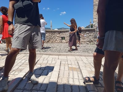 The image shows a sunny day scene with several people near a stone wall in Mostar, Bosnia and Herzegovina. In the foreground, a person wearing grey shorts and white sneakers is partially visible from the waist down. To the left, a woman in a red dress walks away from the camera, and another person in a white t-shirt and blue shorts is visible in the background. In the middle ground, a woman in a dark dress points towards a minaret in the distance, holding a bottle of water. Another woman in a patterned skirt walks behind her. The background features a stone wall, cobblestone ground, a minaret, and a dome, characteristic of the Old Town of Mostar.