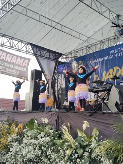 Four young girls dressed in black tops with blue ruffles, rainbow-colored tutus, and headbands perform on an outdoor stage in Sambirejo, Indonesia. The stage is decorated with flowers and foliage. A banner reading "PANAMA ADULNYA SRAGEN, GAMBIREJO, SRAGEN" is visible, alongside another banner with Indonesian text. Several adults are present on the stage, including musicians and observers. Stage lighting and sound equipment are visible.