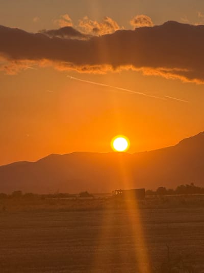 An outdoor vertical shot captures a sunset over a rural landscape in Blatets, Bulgaria. The sun, a bright white orb, is partially obscured by a mountain range in the mid-ground, casting an orange glow across the sky. Dark, textured clouds stretch across the upper portion of the frame, with thin, white contrails visible in the sky. Below the mountains, a field is visible, with a dark structure and a tractor in the distance. The scene is characterized by the warm hues of the sunset and the silhouetted forms of the natural and man-made elements.