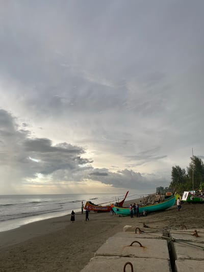 This is an image of a beach in Bateetimoh, Indonesia. The sky is overcast with dramatic clouds. The beach is sandy, and the sea is calm with gentle waves. Several colorful fishing boats are pulled up onto the sand. There are a few people scattered on the beach, some standing near the boats and others walking along the shore. In the foreground, there are concrete blocks, possibly part of a sea wall. The overall impression is of a serene, coastal environment, likely in the late afternoon or early morning given the lighting.