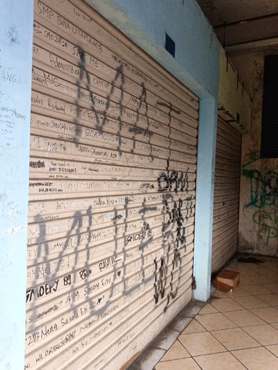 The image depicts a closed storefront in Kidapawan, Philippines, viewed from a low, angled perspective. The primary subject is a cream-colored, corrugated metal roll-down security shutter, extending from floor to ceiling and occupying the central and left portions of the frame. This shutter is extensively covered with numerous handwritten inscriptions in black marker, including names, dates (e.g., "09.02.18," "20/02/2015"), phone numbers/identifiers (e.g., "WA 08xxxxxxxxx," "BM: DD7D8A7"), and short phrases such as "SMP BINA UTAMA," "ADMINISTRATION OFFICER," "Presiden Ridwan," "JANGAN NGAPEZ," "BALIKAN!", "SMOEFY 89," "RAVIN," and "Neng Sayang Egi." A large, stylized black spray-painted tag with angular shapes is prominently overlaid on these inscriptions, casting a distinct shadow.
To the left, a light blue painted concrete pillar or wall segment is visible, also bearing some faint graffiti. Above the shutter, a horizontal light blue painted beam or facade element features a small, dark blue rectangular inset. To the right, another light blue pillar marks a corner, leading into a recessed area. This area contains a second, closed roll-down shutter of similar design, and a concrete wall covered with green and black spray-painted graffiti. A dark rectangular opening, possibly a vent or window, is present in the upper right background, beneath what appears to be red ceiling conduit. The floor is covered with light brown square tiles, showing some dampness or reflections near the base of the main shutter. A small, partially collapsed cardboard box rests on the tiled floor in the lower right corner, against the base of the rightmost pillar. The scene is illuminated by an apparent interior or artificial light source, casting shadows diagonally across the main shutter from the upper left.