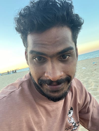 The image shows a man in a mauve-colored sweatshirt taking a selfie on a beach near Kalmunai, Sri Lanka. The background reveals a stretch of beach, the ocean, and a bright sky. A group of people are standing or sitting on the beach in the background. The man has dark hair, a beard, and brown eyes. The sweatshirt has a cartoon character printed on it. The sand is light in color and appears to be wet in some areas.