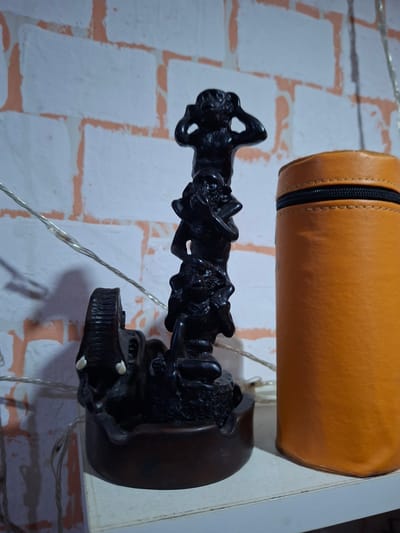 The image shows a close-up view of two objects placed against a wall with a brick pattern. At the top, a cylindrical object made of tan-colored leather is visible. It has a zipper running along its length. Below this, a dark, intricately carved wooden sculpture is positioned. The sculpture appears to depict a group of figures or creatures intertwined. Some of these figures have sharp, tooth-like projections. The background wall has a white base with what looks like orange or reddish-brown mortar lines forming a brick pattern. A thin, silver wire with a twisted texture is also visible in the frame, draping in front of the sculpture and wall. The lighting suggests an indoor setting, possibly artificial. No specific time of day, weather, or emotional cues are discernible. There is no visible text in the image.