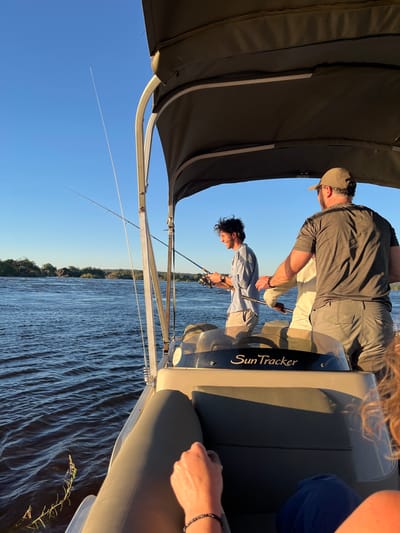 Two men are fishing on a Sun Tracker boat, presumably on the Zambezi River. The location is near Victoria Falls, Zimbabwe. The men are holding fishing rods and looking down at the water. One man wears a gray shirt, khaki pants, and a baseball cap. The other man wears a light blue shirt and khaki shorts. The river is choppy, and the sky is clear blue. Part of the boat's canopy is visible in the top left of the frame.