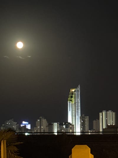 The image depicts a night scene of the Finestrat, Spain skyline. A large, bright full moon is visible in the upper left quadrant of the dark sky, with faint wisps of clouds near it. In the center of the image, a modern, tall skyscraper with distinctive green and yellow lighting accents stands prominently against the night sky. To the right and left of this central tower are several other buildings of varying heights, also illuminated with lights, forming a city skyline. In the foreground, the dark silhouettes of trees and a yellow object, possibly part of a structure or street furniture, are visible, suggesting a viewpoint from ground level or a balcony. The overall impression is a well-lit urban landscape at night, dominated by the bright moon and illuminated architecture.