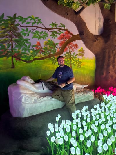 A man stands in a photo booth. The booth is decorated with a painted landscape showing trees and a lawn with green and yellow hues. A white statue is placed in front of the man. He is holding his jacket. A field of illuminated white and green tulips is next to the statue. Above the tulips are a few red flowers. The man is wearing a blue shirt and tan pants. This photo booth is located in Binh Lu, Vietnam.