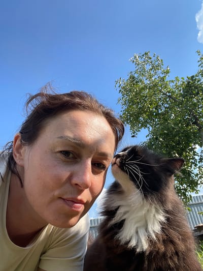 A woman and a black and white cat are captured in a close-up selfie outdoors under a clear blue sky. The woman's face is positioned on the left side of the frame, with her gaze directed towards the camera. Her hair is brown and styled casually, with some strands appearing windblown. The cat is on the right side of the frame, with its face turned towards the woman, gently nudging her nose. The cat has fluffy black and white fur, with prominent white whiskers. In the background, a leafy green tree is visible, its branches reaching towards the sky. A light-colored fence can also be seen behind the subjects. The scene appears to be set in a rural or suburban area, possibly a backyard. The sunlight casts shadows on the woman's face, suggesting it is daytime. The image does not contain any specific indicators to pinpoint the location to Bormist, Russia.