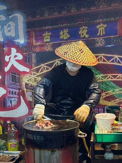 The image shows a person wearing a conical hat and a black jacket, preparing food at a street stall. The person is wearing white gloves and a face mask, and is holding skewers of meat over a large, dark cooking vessel. The background features a vibrant mural with Chinese characters and what appears to be a depiction of a cityscape or industrial scene.
The activity taking place is food preparation, likely for a street food vendor. The skewers of meat suggest a barbecue or grilled dish. The cooking vessel appears to be a large wok or fryer. Various containers and bottles are visible near the stall, including what looks like cooking oil and spices.
The time of day is unclear, but the lighting suggests it could be evening or night, given the artificial lighting and the illuminated background. The weather is not discernible. The location cues, such as the Chinese characters on the mural, indicate an East Asian setting, consistent with the provided location context of Guta, China. The overall mood is functional and perhaps a little gritty, typical of a bustling street food environment.
Visible text includes Chinese characters in yellow and white on the background mural. Some of these characters are repeated and stylized. On a bottle in the foreground, there is yellow text with Chinese characters, appearing to be a brand or product name. The characters are too blurry to be definitively read.