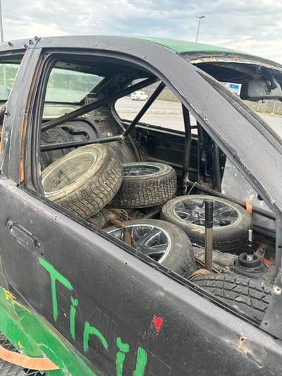 The image shows the interior of a modified car, likely a race car or demolition derby car, viewed from a low angle looking upwards. The car's interior is heavily modified with a roll cage visible throughout. Several tires are stacked inside the car. One tire in the foreground is a light-colored tire with a coarse tread, possibly a snow tire, resting on its side. Behind it, other tires are visible, some with black rims and different tread patterns. The car's body is black and green, with green paint markings visible on the exterior panels, including what appears to be writing in green marker.

The setting appears to be outdoors, with a cloudy sky visible through the open car doors and windows. A lamppost is visible in the background, suggesting an outdoor parking lot or industrial area. The car seems to be positioned in Slitu, Norway, given the context. The overall impression is of a functional but rough vehicle, possibly in the process of being prepared for or having participated in an event. There is no clear activity or event taking place in the image; it is a static shot of the car's interior. The lighting suggests it is daytime.