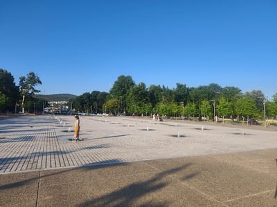 A wide, paved plaza featuring a grid of small water fountains stretches across the foreground and middle ground, under a clear, blue sky. A young girl in an orange skirt and yellow shirt stands near one of the fountains, looking down. In the background, a line of trees borders the plaza, with a cityscape and hills visible beyond. Several people are scattered throughout the plaza, some walking, some sitting on benches. Cars are parked along a road in the far background. The scene appears to be the central plaza of the Thessaloniki Science Center and Technology Museum (NOESIS) in the Thessaloniki Municipal Unit, Greece, likely during daylight hours.