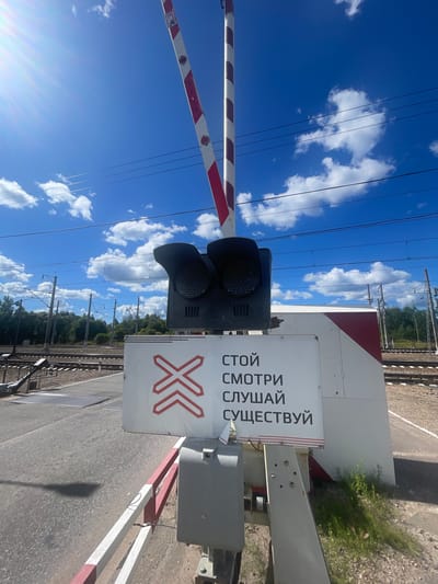 The image depicts a railroad crossing with a barrier arm that is red and white striped and raised. The barrier arm is angled upwards, suggesting it is either in the process of opening or closed. Above the arm, two black signal lights are visible. In front of the barrier, a white sign with red markings is displayed. The sign features a red cross symbol with chevrons pointing inwards on the left side. To the right of the symbol, black text is printed in Russian, which translates to "STOP. LOOK. LISTEN. EXIST." The sign is positioned on a concrete structure, and a small portion of a red and white striped barrier is visible at the bottom of the frame. In the background, railroad tracks extend into the distance, flanked by trees and utility poles with overhead wires. The sky above is a vibrant blue with scattered white clouds, and bright sunlight creates a lens flare in the upper left corner of the image. The overall scene is that of a functioning railroad crossing on a clear day. The location is Kannel'yarvi, Russia.