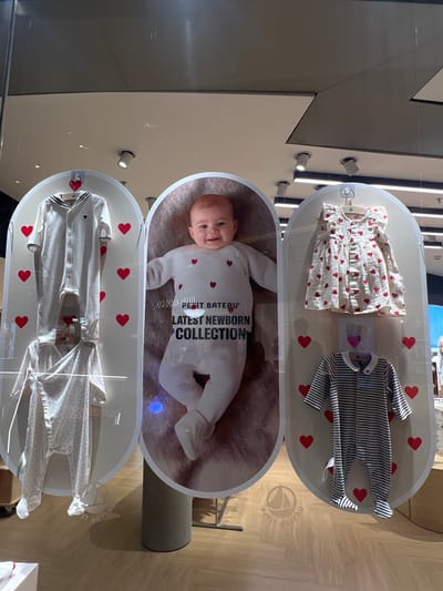 The image shows a vertical display of baby clothing in a retail setting. The display consists of three oval-shaped sections stacked on top of each other. Each section is decorated with small red heart shapes.

The top section contains two baby outfits: one is a white dress with red hearts, and the other is a striped black and white romper.

The middle section contains a photograph of a baby lying on a furry surface. Text reading "Petit Bateau Latest Newborn Collection" is printed on the image.

The bottom section displays two white baby outfits.

The scene appears to be a shop window display, likely in the Nanshan District, China, based on context. The background is blurred, suggesting the display is in a store window facing a street or interior space.