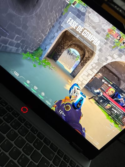 A laptop screen displays a first-person perspective of the game Valorant. The player's hand holds a blue and white pistol. On the screen, the Portuguese text "FASE DE COMPRA" indicates the buy phase. The player's health is at 100. A combat report shows "Rheivenht" with 0 damage caused and 1 kill. The map features stone arches and water. The laptop keyboard is visible, with a power button lit red.