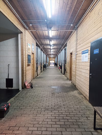 The image shows a long, indoor corridor in Chernogolovka, Russia, possibly a stable or equestrian facility. The corridor has brick flooring, wooden plank walls, and a wooden plank ceiling. Overhead, fluorescent lights are suspended. Along both sides of the corridor are horse stalls with open tops and metal bars. Some stalls are occupied by horses. A black toolbox, pink storage container, and bench are placed along the corridor. An "INFORMATION" sign is mounted on the wall.