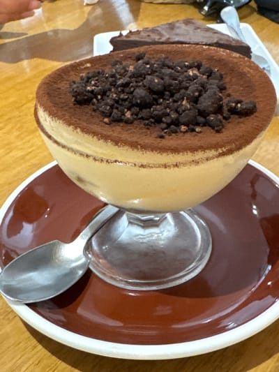 A Tiramisu dessert sits on a dark brown saucer with a white rim, accompanied by a silver spoon. The dessert appears to be in a glass, tilted slightly. A portion of chocolate cake sits on a white plate to the left of the glass. This scene is located in Thong Sala, Thailand.