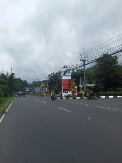 A straight road is lined with trees and foliage. A Pertamina gas station sign displays fuel prices. Several motorbikes are on the road. A utility pole with wires is visible. The sky is overcast with gray clouds. The scene appears to be in a rural or suburban setting, potentially near a mountainous area.